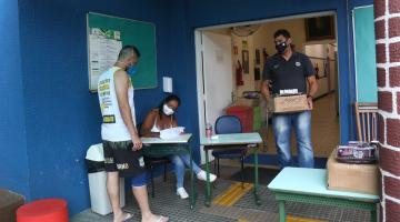 Homem está carregando um caixa, na passagem entre uma sala para outra em escola. Outro homem, à esquerda, está sendo atendido por uma mulher que mexe em papeis. #Paratodosverem