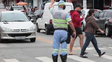 agente de trânsito está parado sobre faixa sinalizando para veículos com a mão esquerda para o alto. Ele está de costas para a foto. Dois pedestres atravessam. #paratodosverem