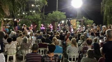 público está sentado em praça ao ar livre. é noite. Uma forte luz ilumina o ambiente. Todos assistem a um show. #paratodosverem