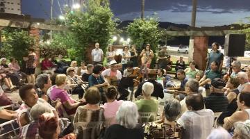 pessoas estão sentadas em cadeiras no meio de praça assistindo apresentação musical. #paratodosverem