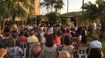 várias pessoas sentadas e em pé numa praça assistindo a um show ao fundo no fim de tarde. #paratodosverem