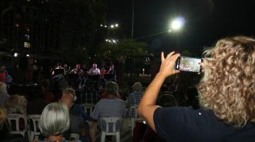 noite de apresentação musical, com músicos ao fundo. Há pessoas sentadas na plateia. Em primeiro plano, à direita, uma mulher de costas para a foto, aponta o celular para fotografar a banda. #paratodosverem