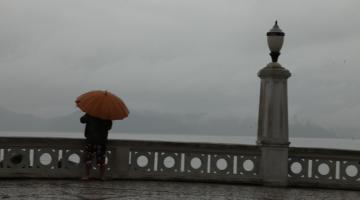 Pessoa está passada atrás de mureta na Ponta da Praia em um dia chuvoso. Ela segura um guarda-chuva aberto. #Paratodosverem