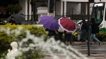 pessoas caminham em rua com guarda-chuva. em primeiro plano aparecem ás águas de um chafariz e um arbusto. #paratodosverem 