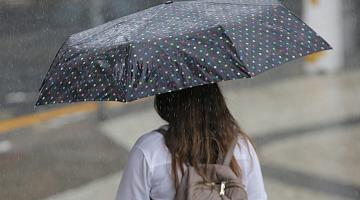 Mulher está de costas para a foto. Ela segura um guarda-chuva na rua. Está chovendo. #Paratodosverem