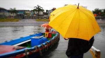 pessoa está na estação de catraias parada do lado direito segurando um guarda-chuva. Ela está de costas. Ao lado esquerdo está chegando uma catraia com uma pessoa dentro. O dia está nublado. #paratodosverem 