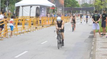 Ciclista pedala pela avenida da orla durante competição com outros atletas ao fundo. #pratodosverem