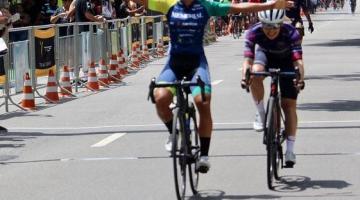 Ciclista montada na bicicleta está com os dois braços no ar, comemorando a chegada em primeiro lugar. Ela está na pista. Atrás vem outra atleta. #paratodosverem