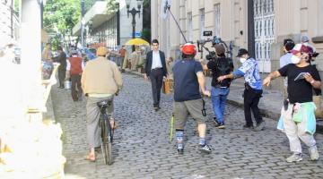 várias pessoas se movimentando em rua do centro para fazer uma filmagem. Alguns seguram equipamentos como câmeras e microfones. Outros estão com roupas antigas. #paratodosverem