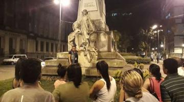Várias pessoas estão em torno da estátua de Brás Cubas. Uma mulher orienta o grupo. #Paratodosverem