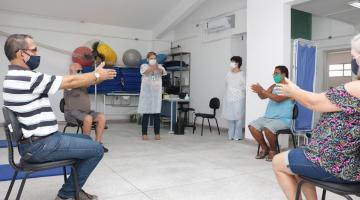 Pessoas estão sentadas em cadeiras fazendo movimento com os braços. Uma orientadora está em pé. Ao fundo, há material para exercícios, como colchonetes e bolas. #Paratodosverem