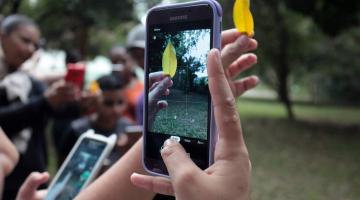 imagem em close de mão segurando um celular que está filmando uma folha na ponta dos dedos de outra mão. #paratodosverem