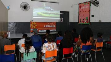 jovens estão sentados em cadeiras, de costas para a foto, assistindo a uma projeção num telão na parede. #paratodosverem