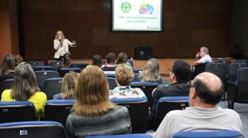 várias pessoas sentadas em auditório. Uma pessoa fala ao fundo. Ela está à frente de uma projeção onde aparece o símbolo da Cipa, algo escrito e mais uma arte. #paratodosverem