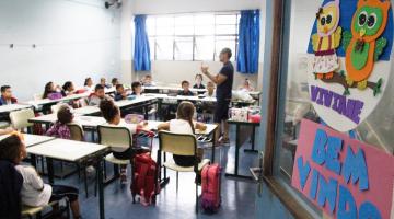 Porta de escola está aberta. Ao fundo o professor e os alunos sentados. #Paratodosverem