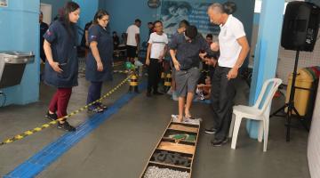 alunos fazem atividade sensorial pisando em uma espécie de brinquedo no chão com várias texturas.Monitores acompanham. #paratodosverem