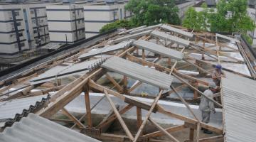 Homens colocam telhas na cobertura do bloco 1 de escola. Ao fundo, os prédios de um conjunto habitacional. #paratodosverem