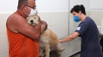 Homem está em pé abraçado a uma cachorro em atendimento veterinário. Outro homem toca nas costas do animal. #Paratodosverem