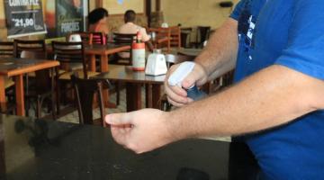 Homem borrifa álcool em uma das mãos. Ao fundo estão mesas de um restaurante. #paratodosverem