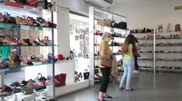 mulheres andam dentro de loja de calçados. Há várias prateleiras. .#Paratodosverem
