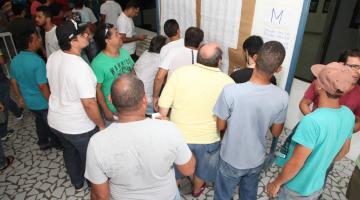 candidatos olham folhas em painel de madeira #paratodosverem