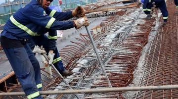 Homens uniformizados concretam viaduto. Eles estão em meio a ferragens. #Pracegover