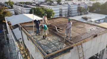 homens estão na parte superior de uma estrutura no alto de um prédio. Eles lançam e assentam concreto sobre armação de ferro. #paratodosverem