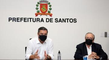 o prefeito Rogério ao lado de um homem. Eles estão sentado. Há um brasão da Cidade afixado na parede atrás e abaixo se lê Prefeitura de Santos. #paraotodosverem