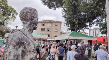 Busto de Zumbi dos Palmares, de máscara, com público em volta acompanhando cerimônia. #pracegover