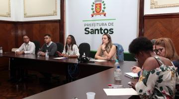 reunião com sete pessoas sentadas em torno de uma mesa. Ao fundo, um banner com o brasão da prefeitura. #paratodosverem