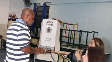Homem se dirige a cabine de votação conversando com mesárias. #pracegover