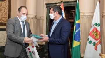 O cônsul à esquerda e o prefeito à direita. Ambos estão em pé, trocando presentes, à frente das bandeiras de São Paulo, Brasil e Santos. #paratodosverem