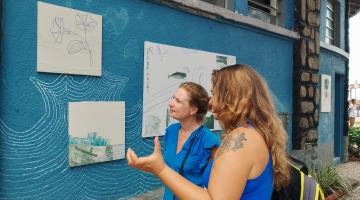 duas mulheres estão observando quadros em muro do aquário. #paratodosverem 