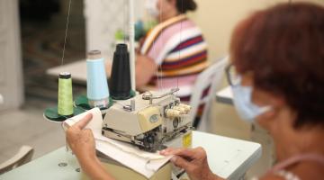 duas mulheres costurando em máquina. O foco está no primeiro plano.#paratodosverem