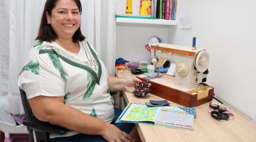 Alessandra está sentada diante de bancada onde está máquina de costura. Ela sorri para a foto. #paratodosverem