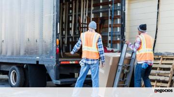 Dois homens carregam mercadoria para dentro de caminhão. #paratodosverem