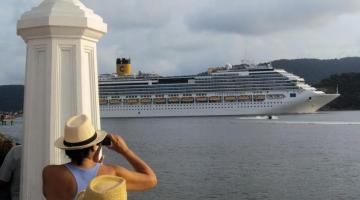 Homem de costa, com chapéu, observa a passagem de navio de cruzeiros pelo estuário de Santos. #paratodosverem