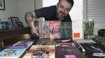 homem está atrás de mesa com diversos livros. Ele aponta com a mão direita para as publicações e sorri. #paratodosverem