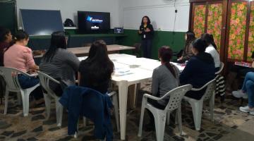 sala com mesa ao centro e várias mulheres sentadas. Uma está em pé falando ao grupo. Ela está ao lado de um monitor. #paratodosverem