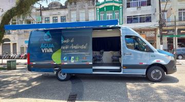 van parada em praça com porta aberta e toldo na frente. Exterior do veículo é estampado com logotipo do programa água viva, simbolizado por uma grande gota d'água. #paratodosverem