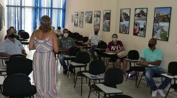 sala com alunos adultos sentados. Todos usam máscaras e estão afastados. Professora está de costas para a foto, em pé. #paratodosverem