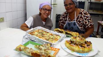 pizzas de forno estão expostas sobre mesa. Duas mulheres estão atrás sorrindo. #paratodosverem