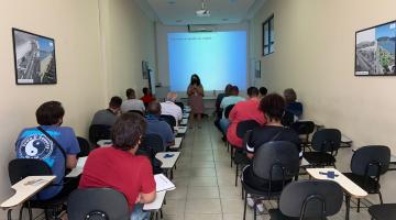 alunos do curso estão sentados de costas para a foto, assistindo à aula. Há uma mulher falando ao fundo. Ela está diante de um telão. #paratodosverem