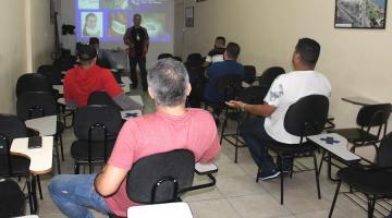 pessoas estão sentadas em sala de aula. Ao fundo um homem fala ao grupo. Ele está diante de uma projeção na parede sobre imobilização cervical. #paratodosverem