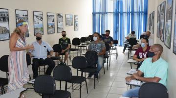 Sala de aula com pessoas sentadas em cadeiras espaçadas entre si. Mulher que está em pé fala ao grupo. #Paratodosverem