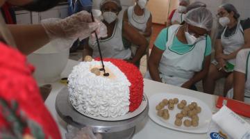 close em mãos que formam o rosto do papai noel sobre o chantilly do bolo. Alunas estão ao fundo observando e fazendo anotações. #paratodosverem