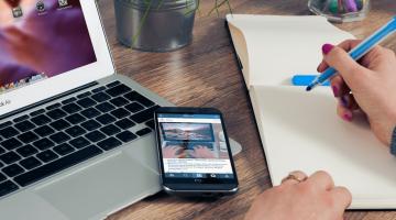 notebook aberto, smarthphone em cima e mãos de mulher fazendo anotação em papel. #paratodosverem