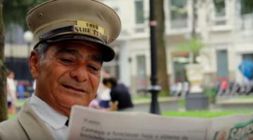 Motorneiro de bonde lendo jornal em praça. Imagem para Santos Eu te Amo. #paratodosverem