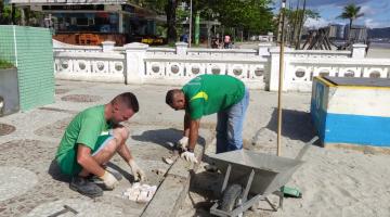 dois homens uniformizados fazem reparos em mosaico português no calçadão interno da orla. #paratodosverem 