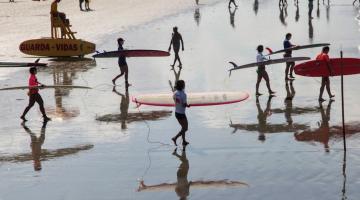 surfistas carregam prancha na beira d'água. #paratodosverem 